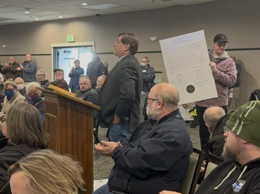 Troy speaks at lectern while person holds giant copy of a joint resolution up behind him